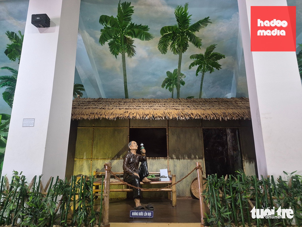 Gian nhà lá trong thời chiến với hàng cau, bụi chuối và lu vại quanh nhà. Giữa không gian ấy là hình ảnh người mẹ già, tay cầm ngọn đèn dầu do mẹ tự chế, báo hiệu cho du kích nhận biết có địch - Ảnh: TRẦN THƯỜNG